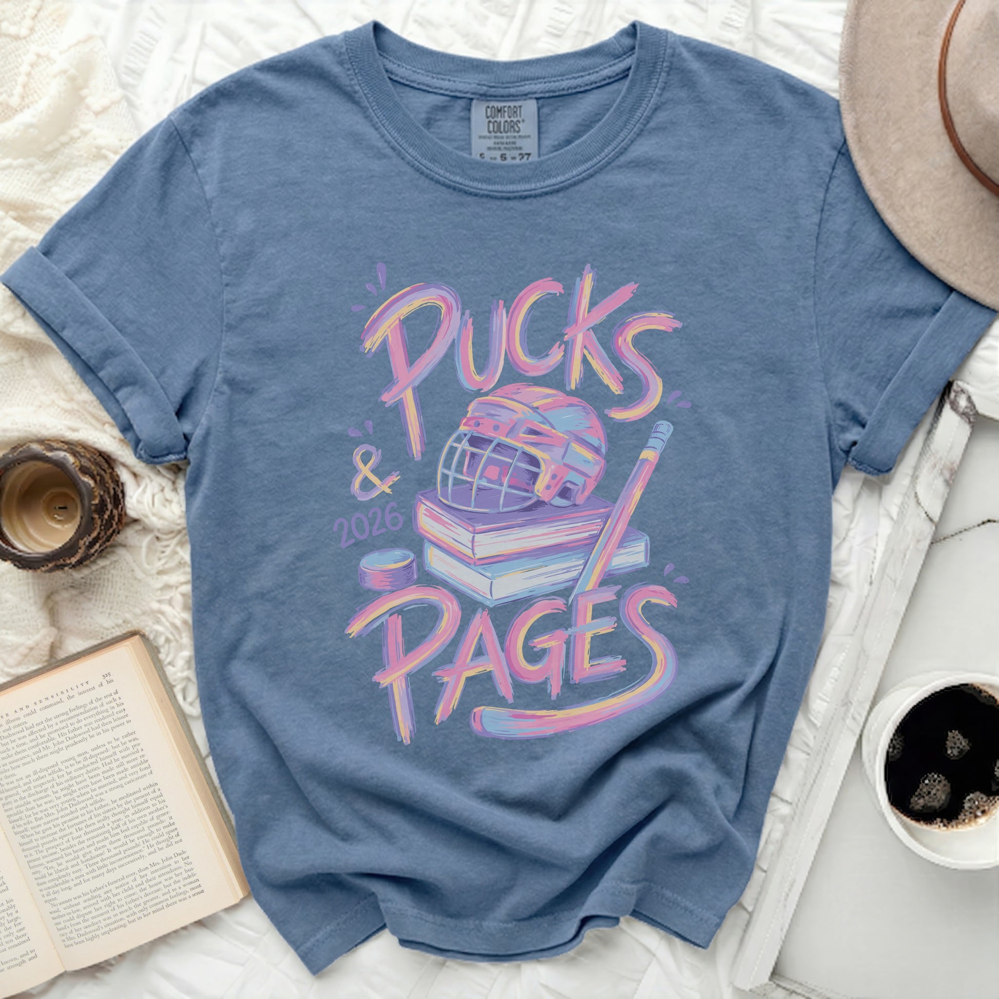 Blue t-shirt with "Pucks & Pages 2026" in colorful text and hockey gear.