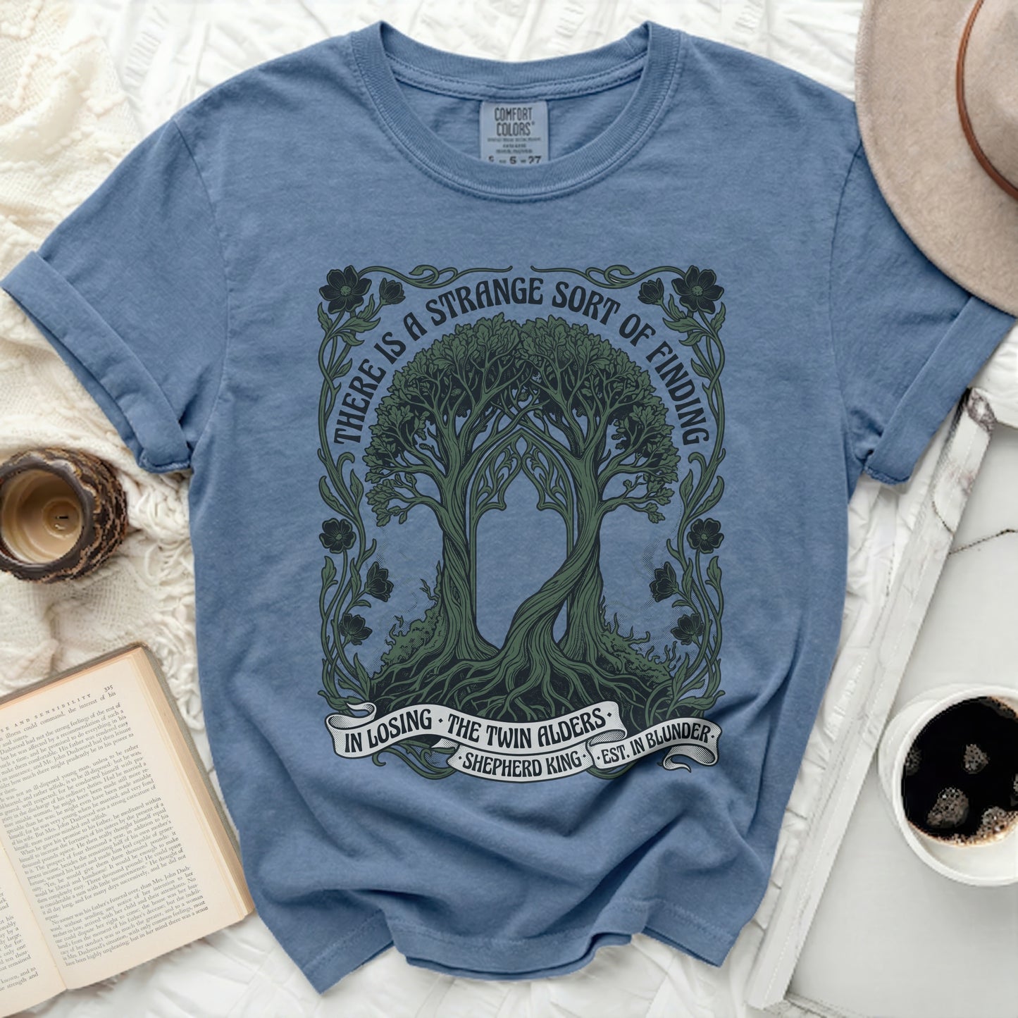 Blue t-shirt with trees and text "There is a strange sort of finding in losing the twin alders."