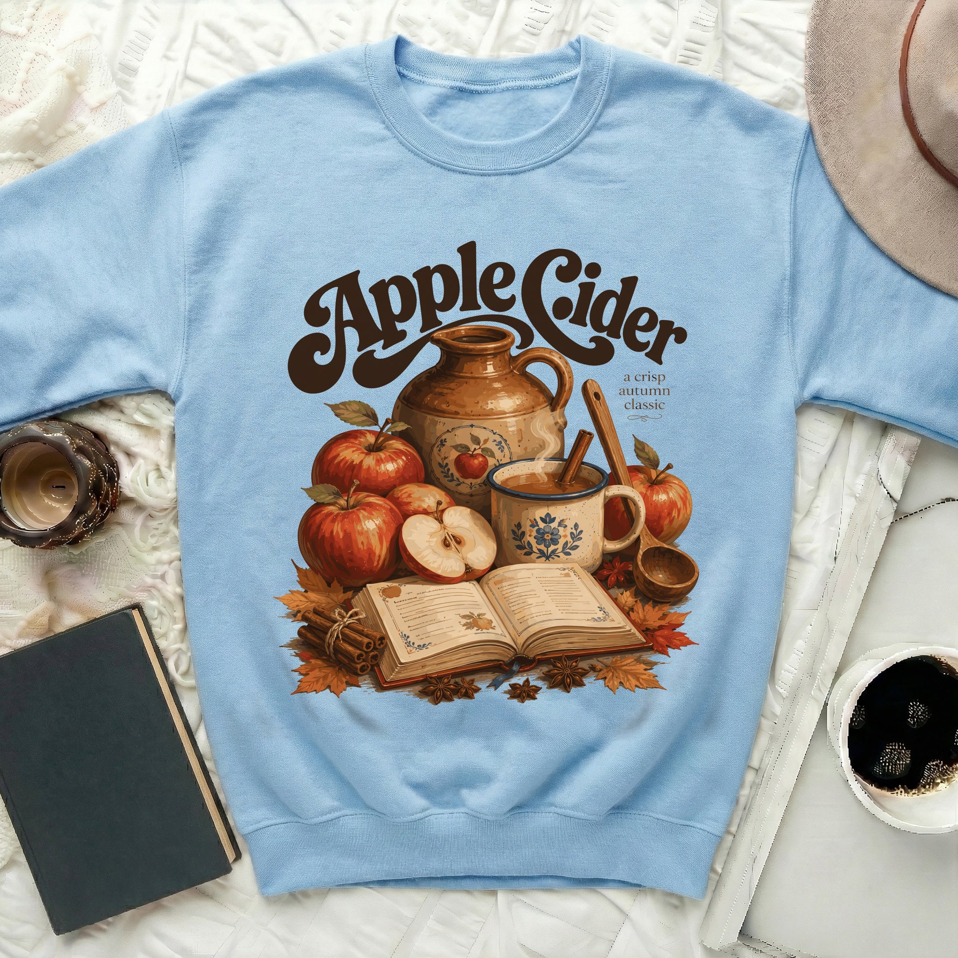 Light blue crewneck sweatshirt with "Apple Cider" graphic featuring apples, warm drinks, and autumn leaves.