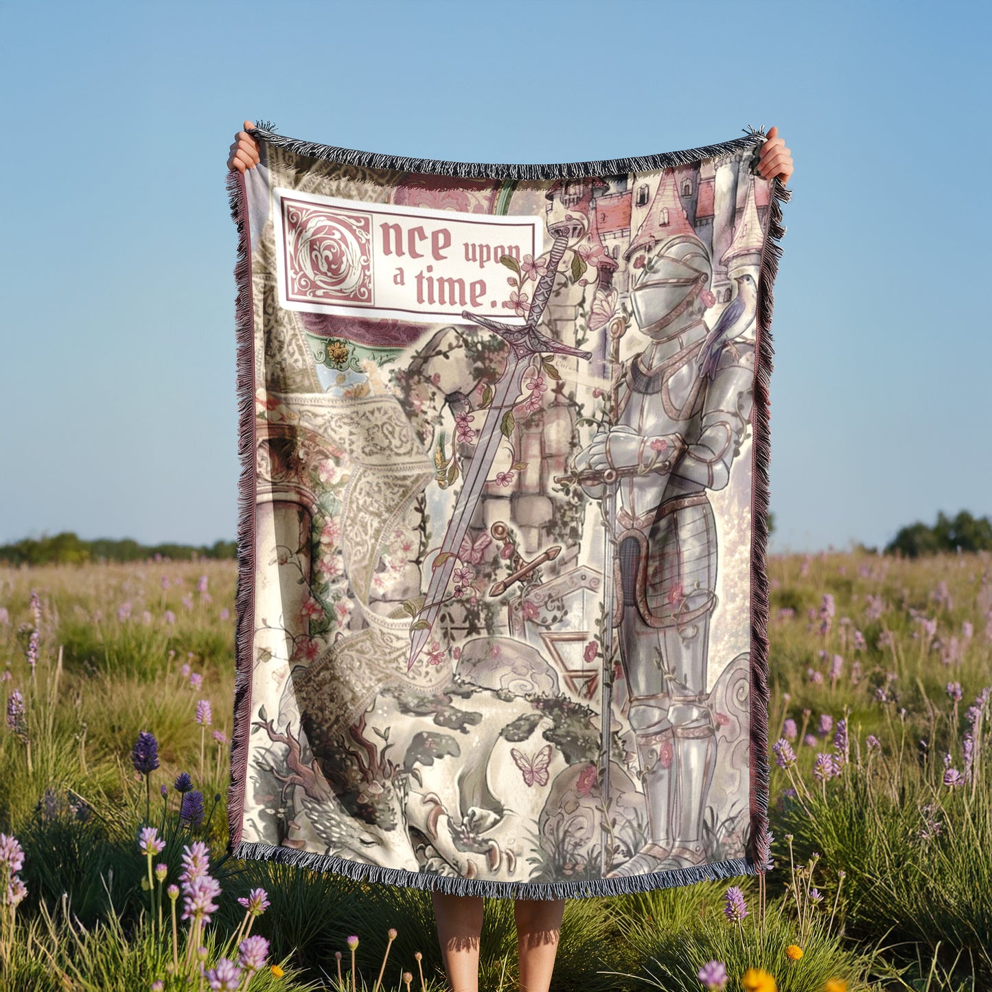 A person is holding up a large, colorful blanket with a medieval-style illustration on it, standing in a field of purple flowers.