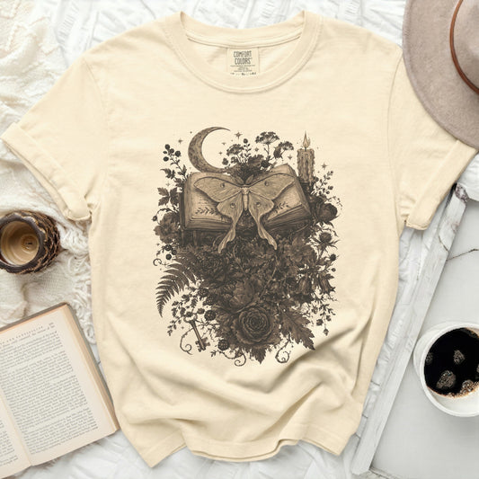 Cream t-shirt with a sepia-toned moth on an open book surrounded by flowers.