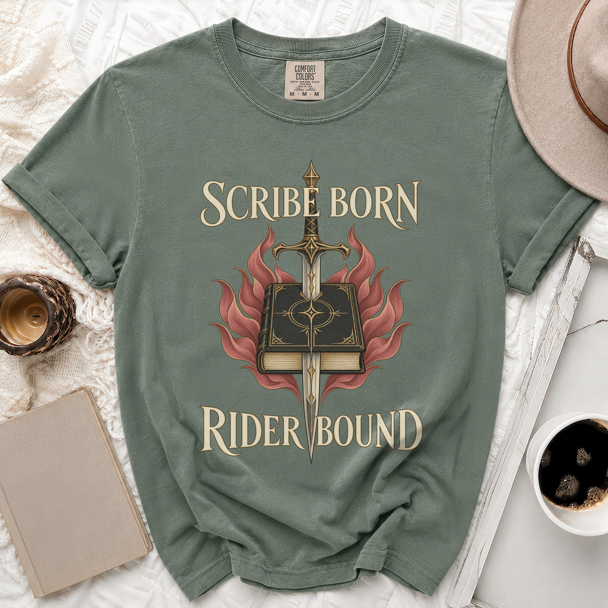 A muted green t-shirt with a graphic of a book pierced by a sword, surrounded by red flames, and the text Scribe Born Rider Bound.