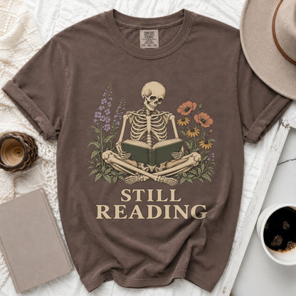 Brown t-shirt with skeleton reading book design and text "STILL READING".