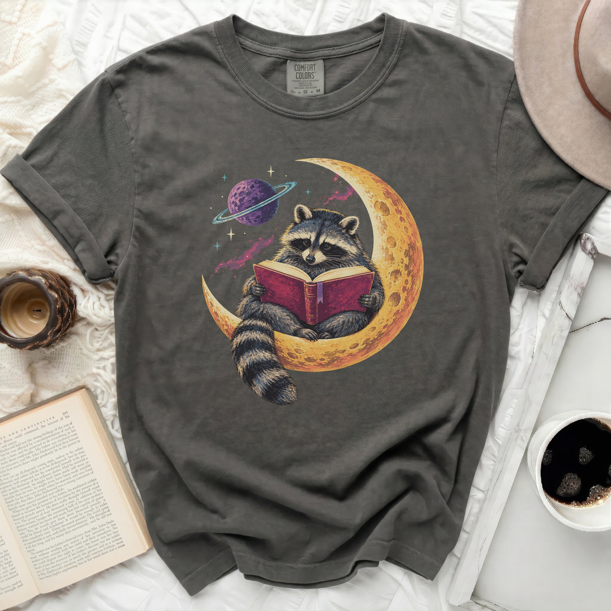 Raccoon reading a book on a crescent moon with a planet and stars.