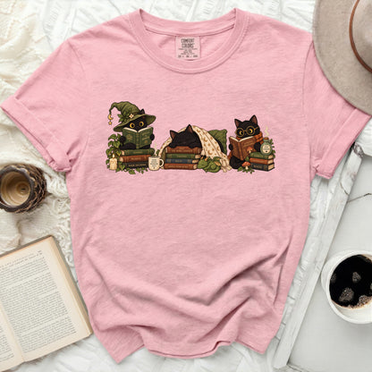 Pink t-shirt with illustration of three black cats reading books and wearing wizard hats.