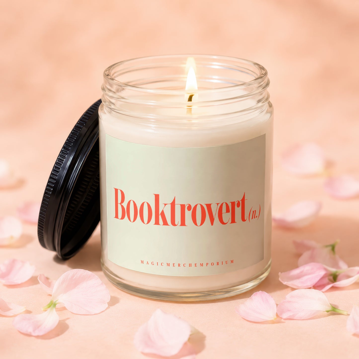 Lit candle in glass jar with "Booktrovert (n.)" label, petals on pink background.