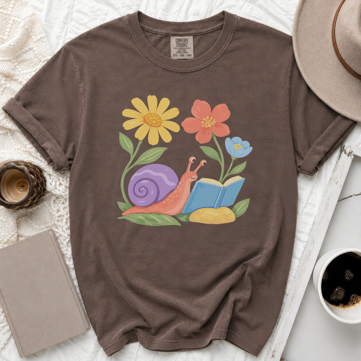 Brown t-shirt with a graphic of a snail reading a book among flowers.