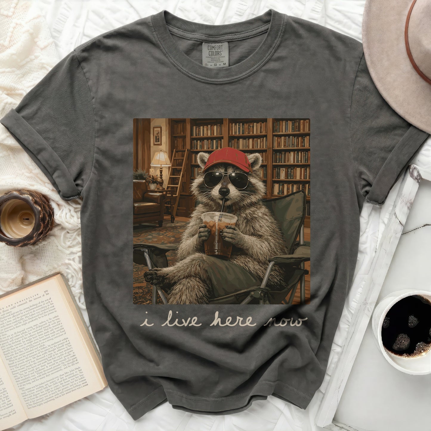 Raccoon wearing sunglasses and a red hat drinks iced coffee, text "i live here now".