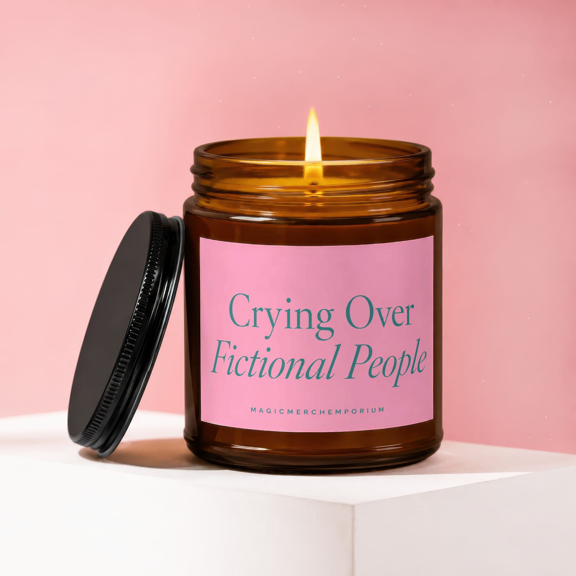 Amber candle with lit wick, black lid, and pink label reading "Crying Over Fictional People".