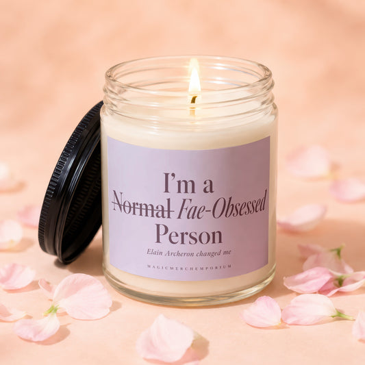 A lit candle in a clear glass jar with a purple label reading "I'm a Normal Fae-Obsessed Person" surrounded by scattered pink flower petals.