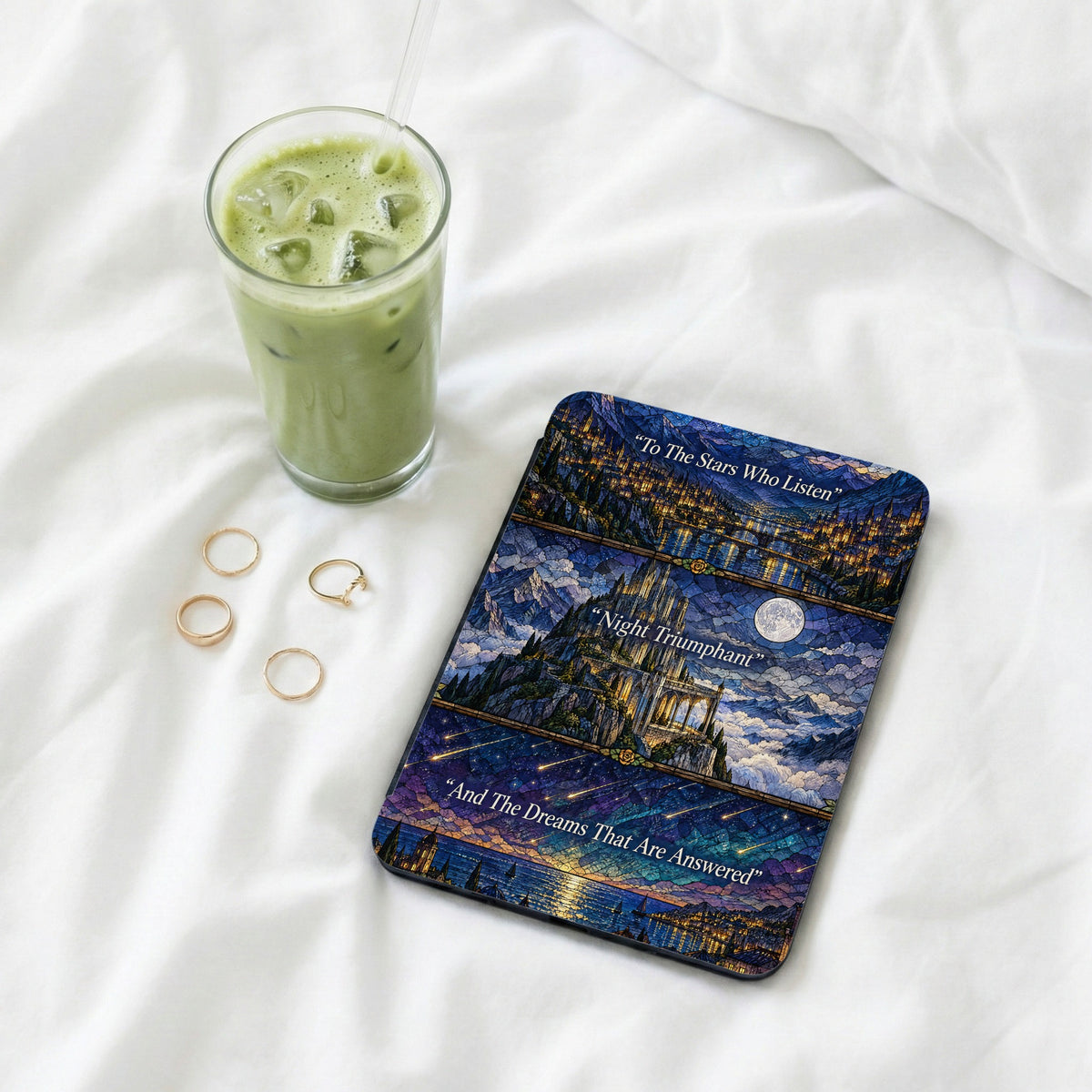 An e-reader with a stained glass style fantasy castle cover rests on white bedsheets beside a glass of iced matcha.