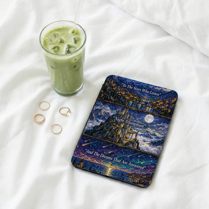 An e-reader with a stained glass style fantasy castle cover rests on white bedsheets beside a glass of iced matcha.