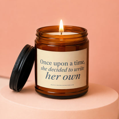 Amber glass candle with lit wick and text "Once upon a time, she decided to write her own."