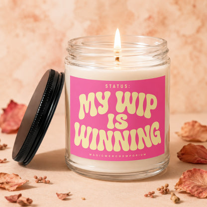 Lit candle in glass jar with pink label reading "STATUS: MY WIP IS WINNING"