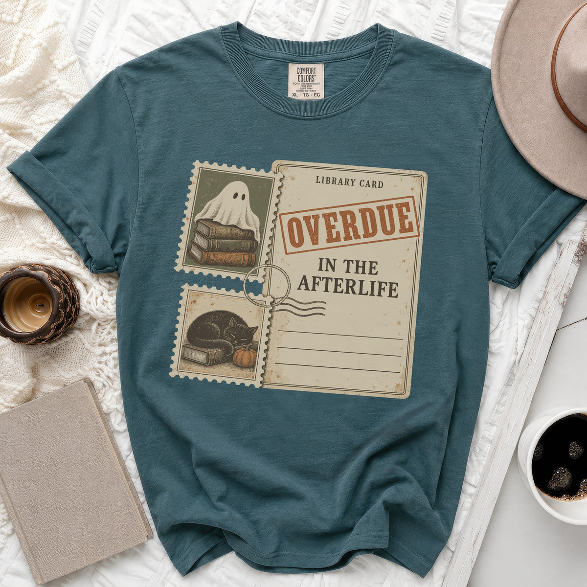 Teal t-shirt with a graphic of a library card and stamps featuring a ghost, books, cat, and pumpkin.