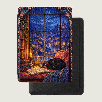 A closed black e-reader features a colorful cover showing a stained glass window with a rainy city view, books, a candle, and a sleeping cat.