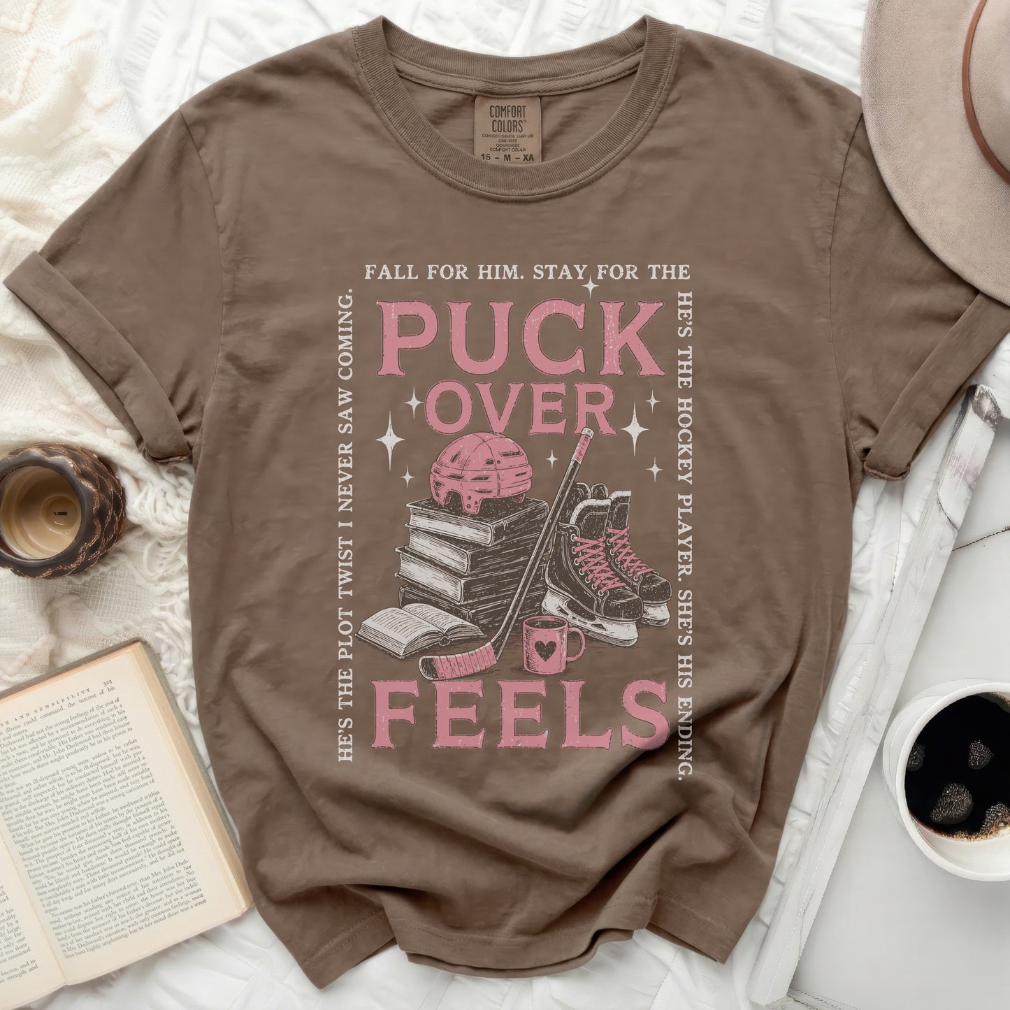 Brown t-shirt with "Puck Over Feels" text, hockey gear, and stacked books.
