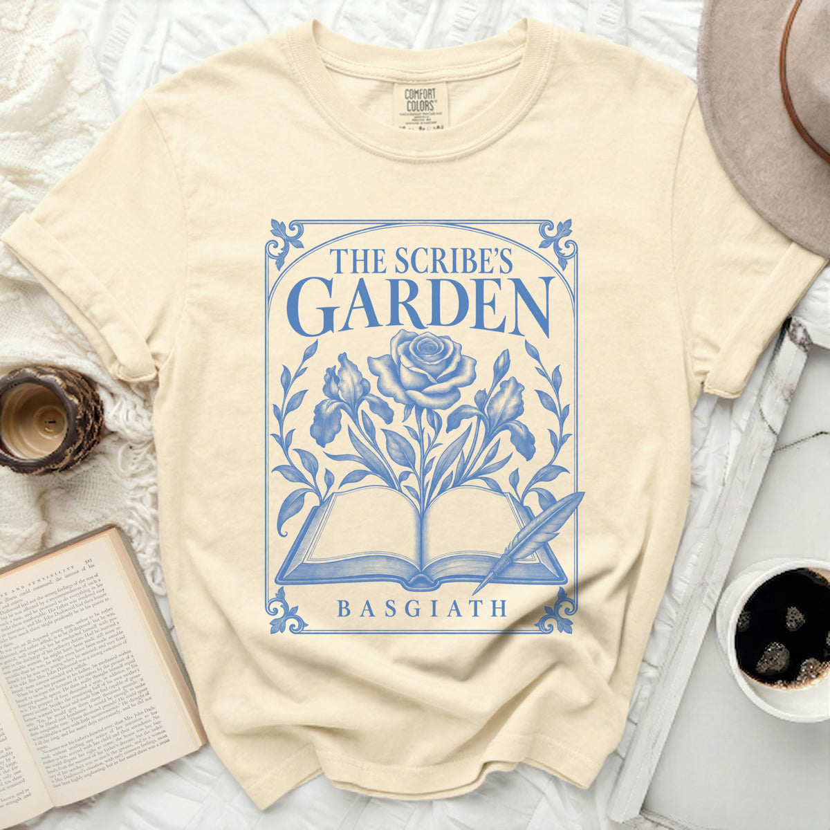 Cream t-shirt with blue graphic of open book, rose, iris, and feather, reading "THE SCRIBE'S GARDEN BASGIATH".