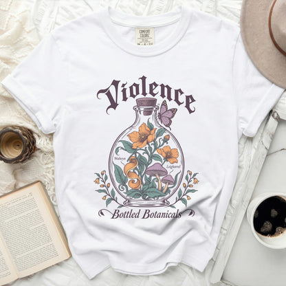 White t-shirt with a graphic of a glass bottle containing flowers, mushrooms, and a butterfly, with text reading "Violence Bottled Botanicals".