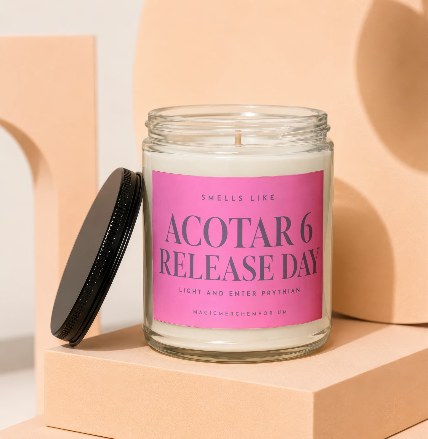A scented candle in a glass jar with a black lid and pink label reading Acotar 6 Release Day.