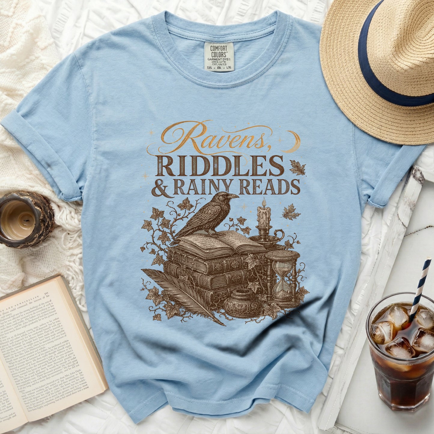 Light blue t-shirt with gold text "Ravens, Riddles & Rainy Reads" and vintage illustration.
