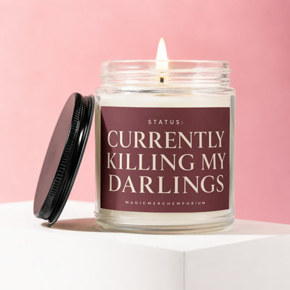 Glass jar candle with a lit wick and a maroon label that reads "Status: Currently Killing My Darlings."