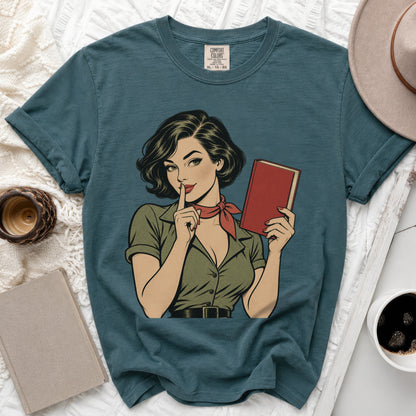 Comic book style graphic of woman holding book and gesturing for quiet on teal t-shirt.