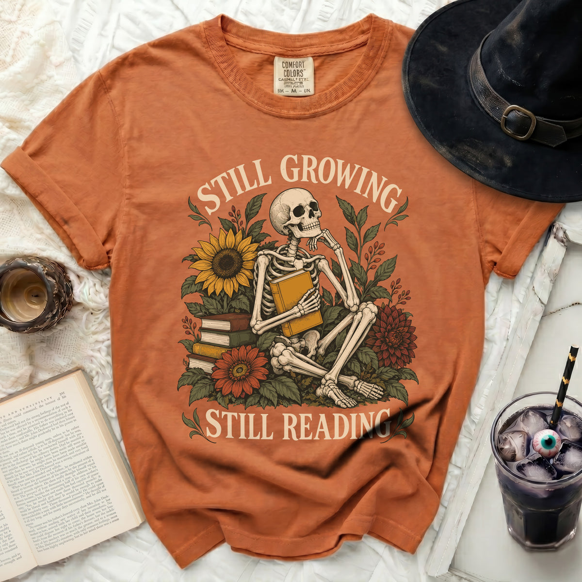 Orange t-shirt with skeleton reading among flowers, "Still Growing Still Reading" text.