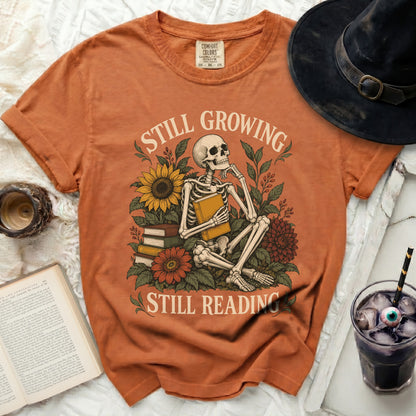Orange t-shirt with skeleton reading among flowers, "Still Growing Still Reading" text.