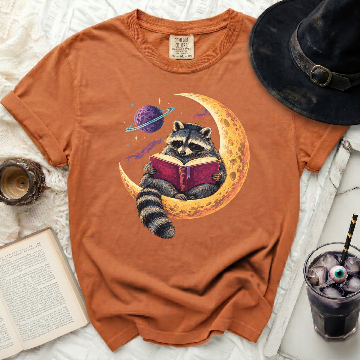 Orange t-shirt with a raccoon reading a book on the moon and a planet.