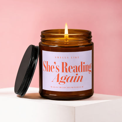Amber jar candle with lit wick, reads "She's Reading Again" and "MagicMerchEmporium".