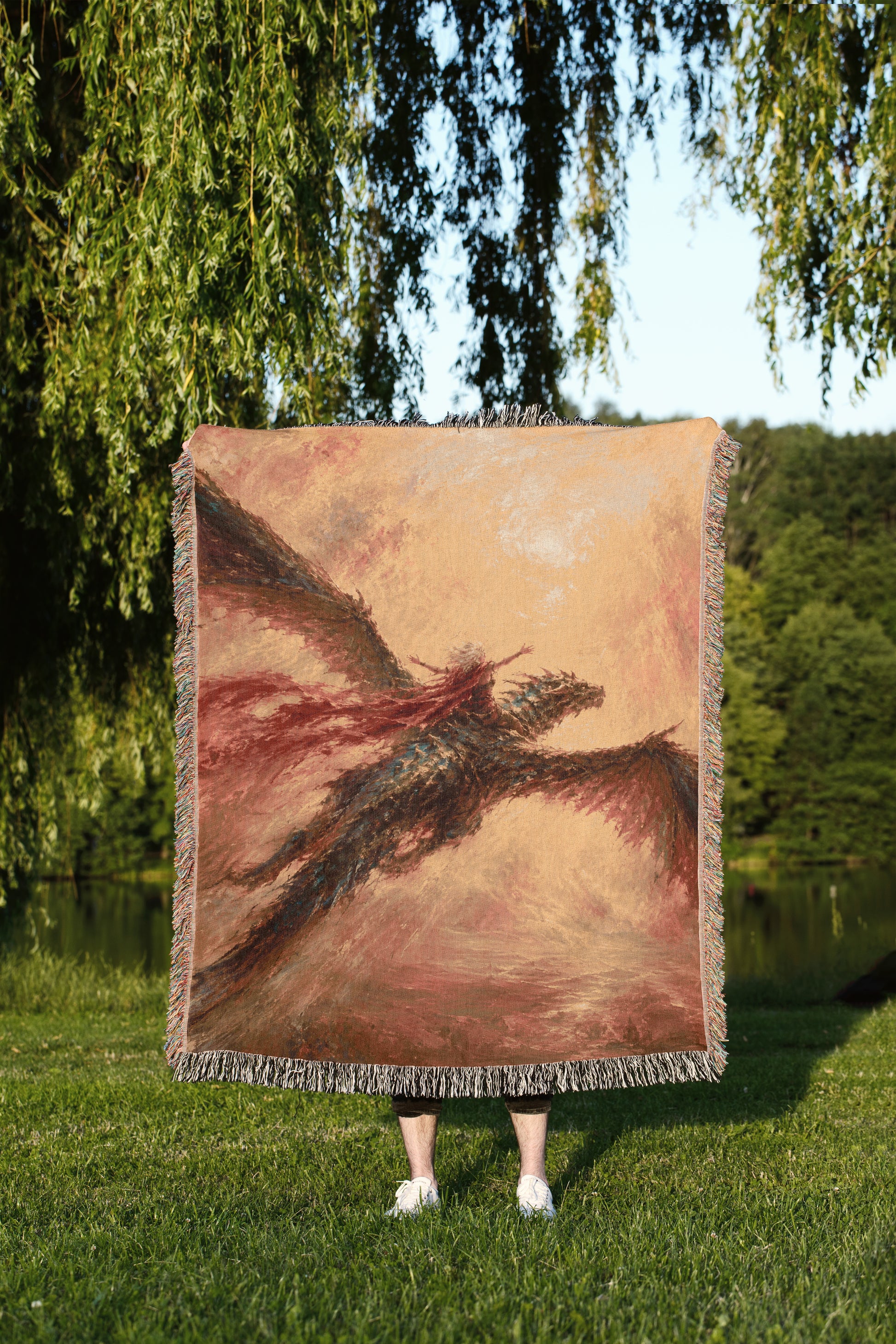 A person is standing in a grassy area, holding a large square tapestry or blanket that features a painting of a dragon or mythical creature.