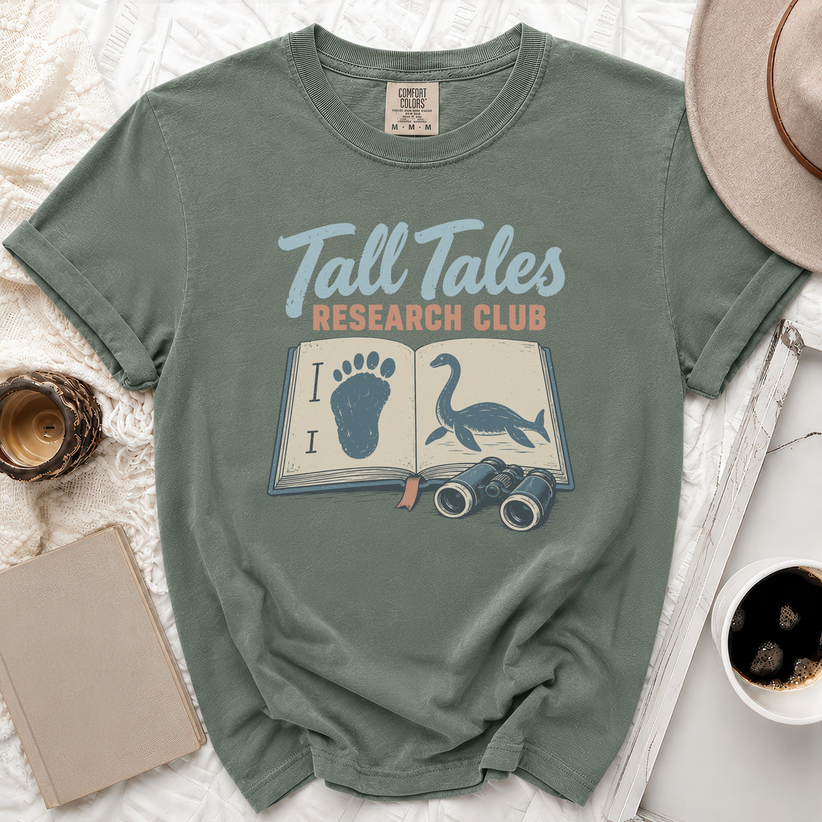 Green t-shirt with "Tall Tales Research Club" text and open book with footprints and Loch Ness Monster.