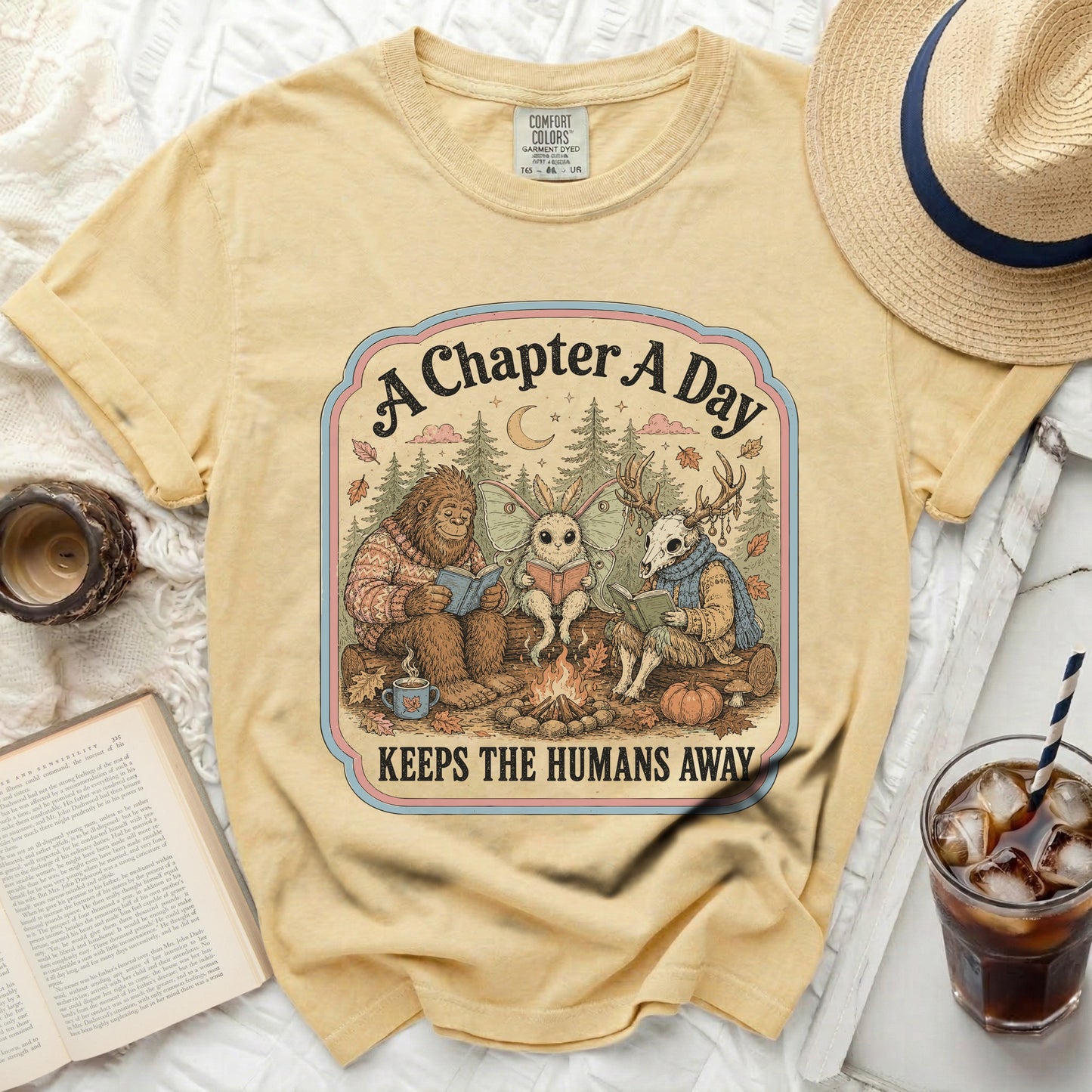 Yellow t-shirt with Bigfoot, moth fairy, and deer skull reading books by campfire.