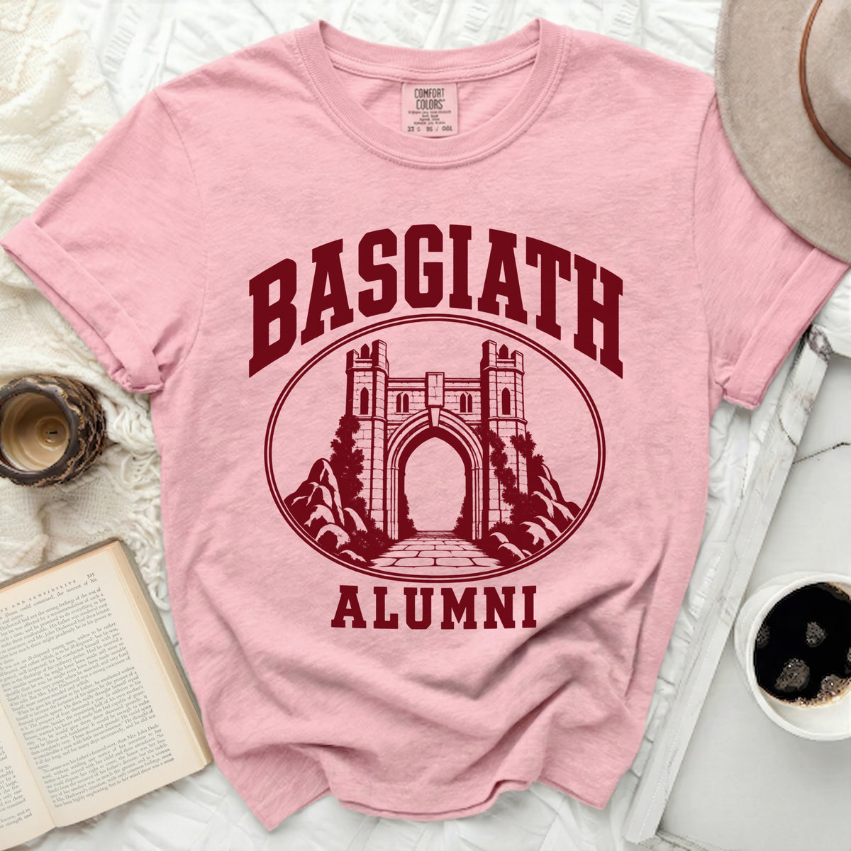 A pink t-shirt featuring a maroon graphic of a stone archway, with the text Basgiath Alumni.