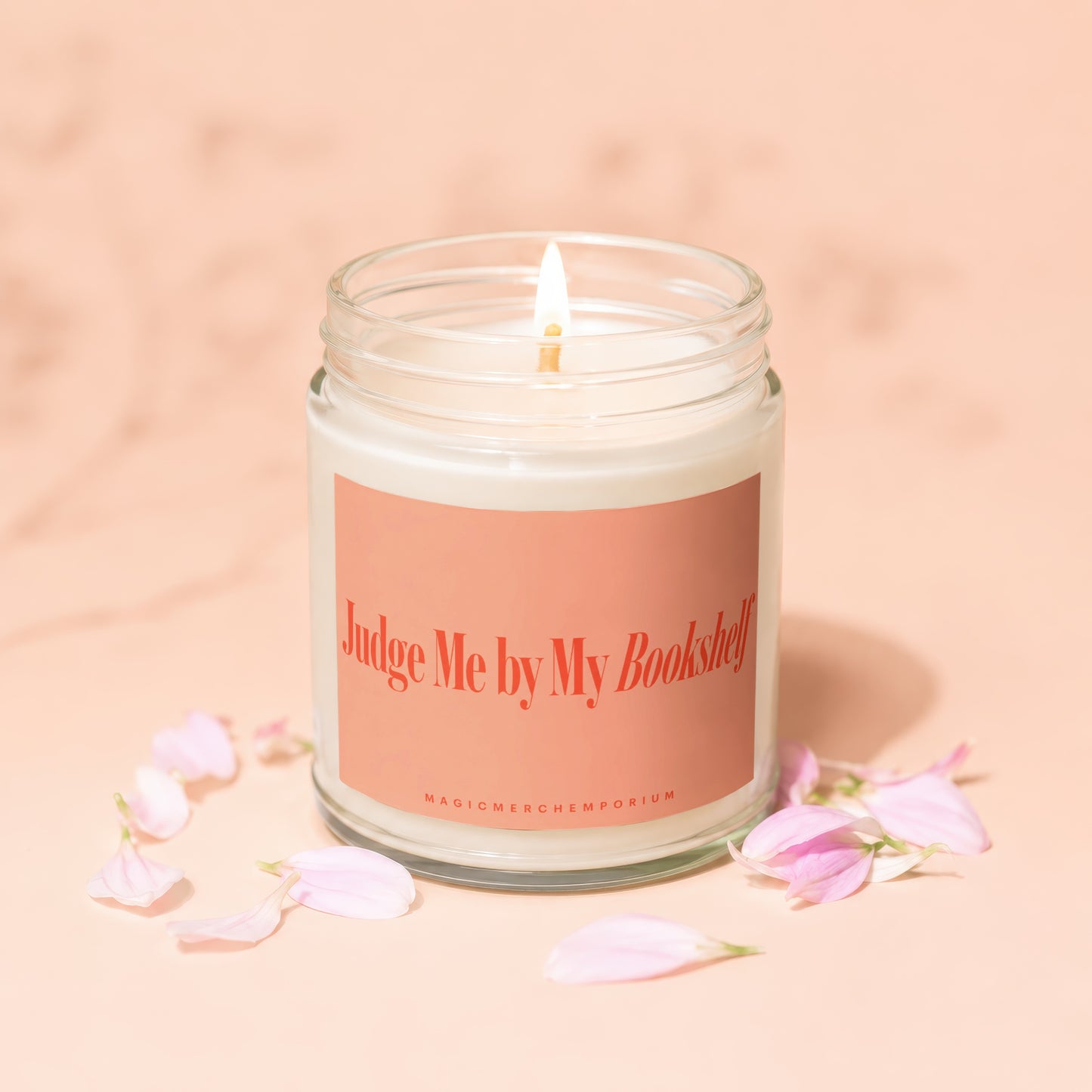 Lit candle in glass jar with "Judge Me by My Bookshelf" label and pink petals.