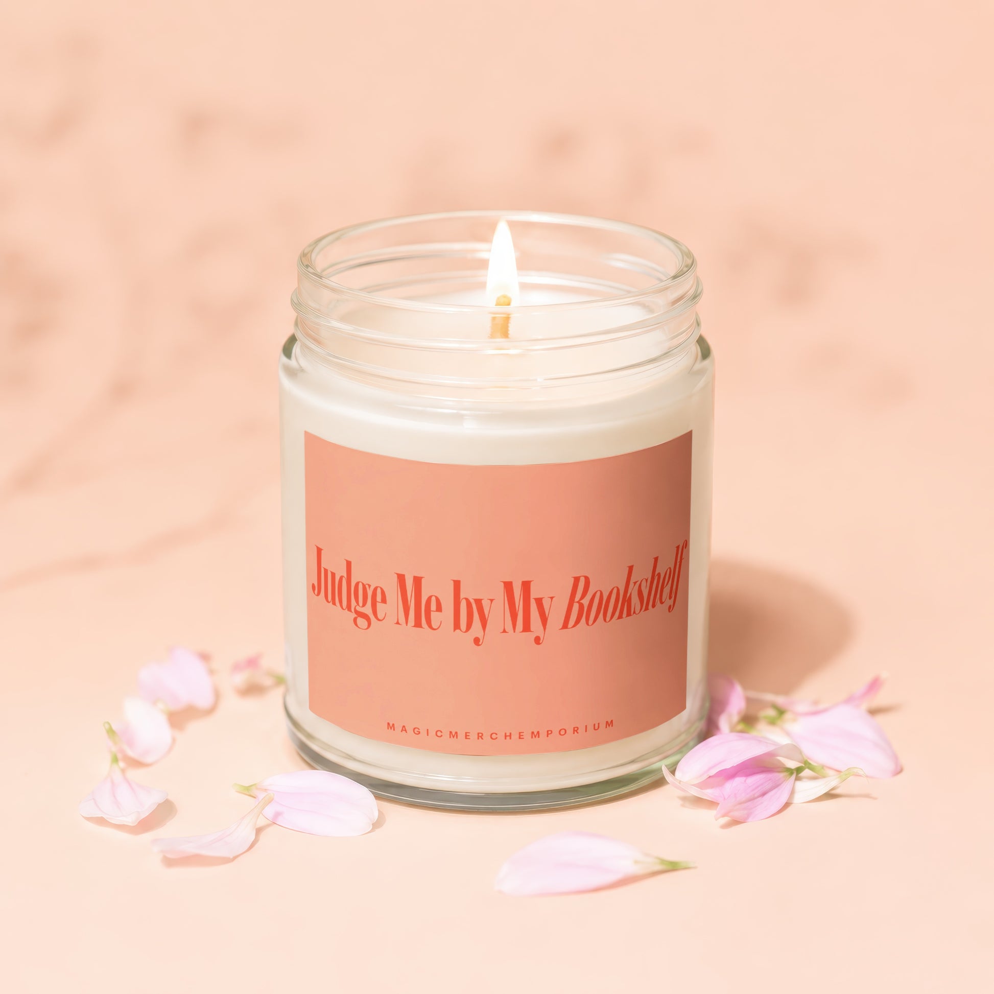 Lit candle in glass jar with "Judge Me by My Bookshelf" label and pink petals.