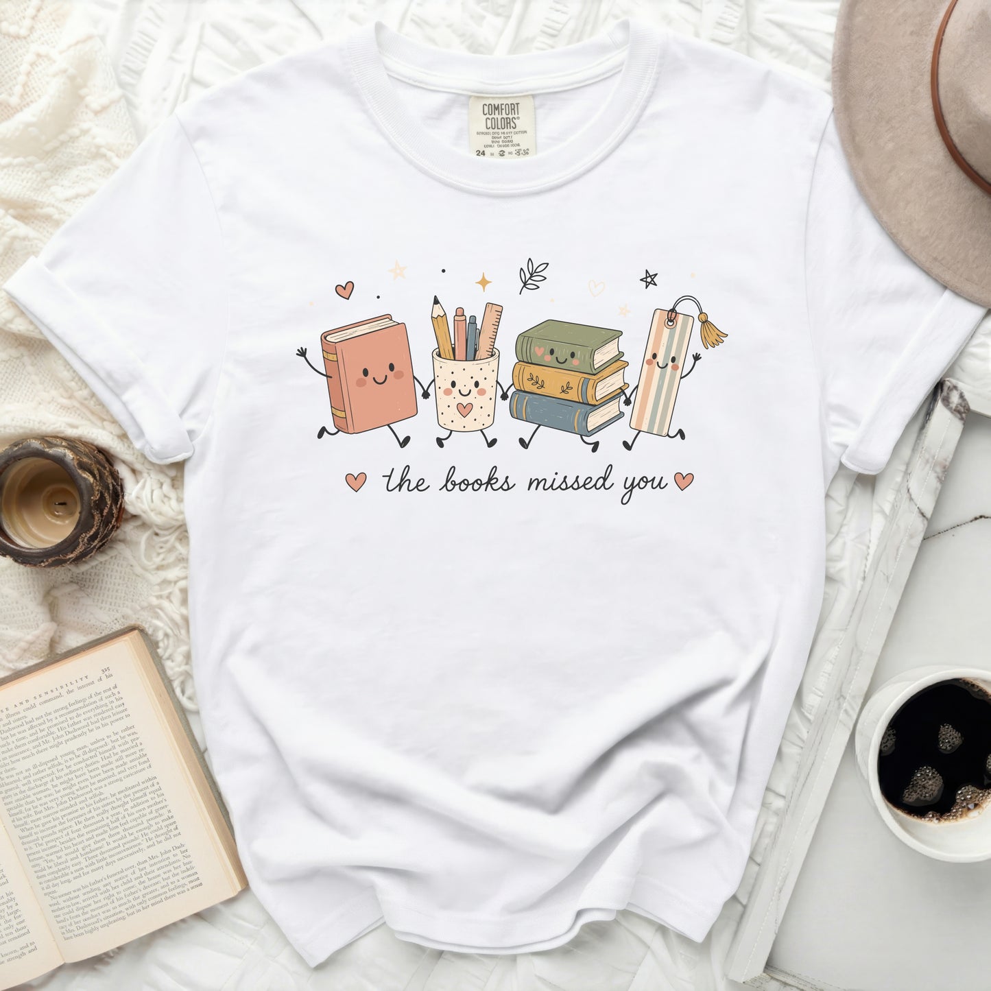White t-shirt with graphic of happy books and school supplies characters saying "the books missed you".