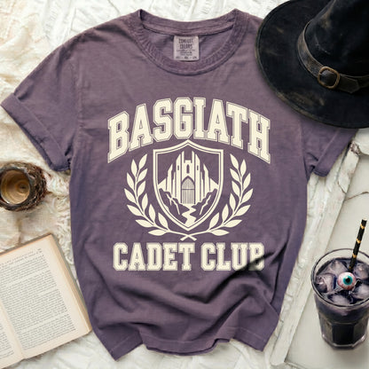 Purple t-shirt with "Basgiath Cadet Club" text and crest, next to a hat and drink.
