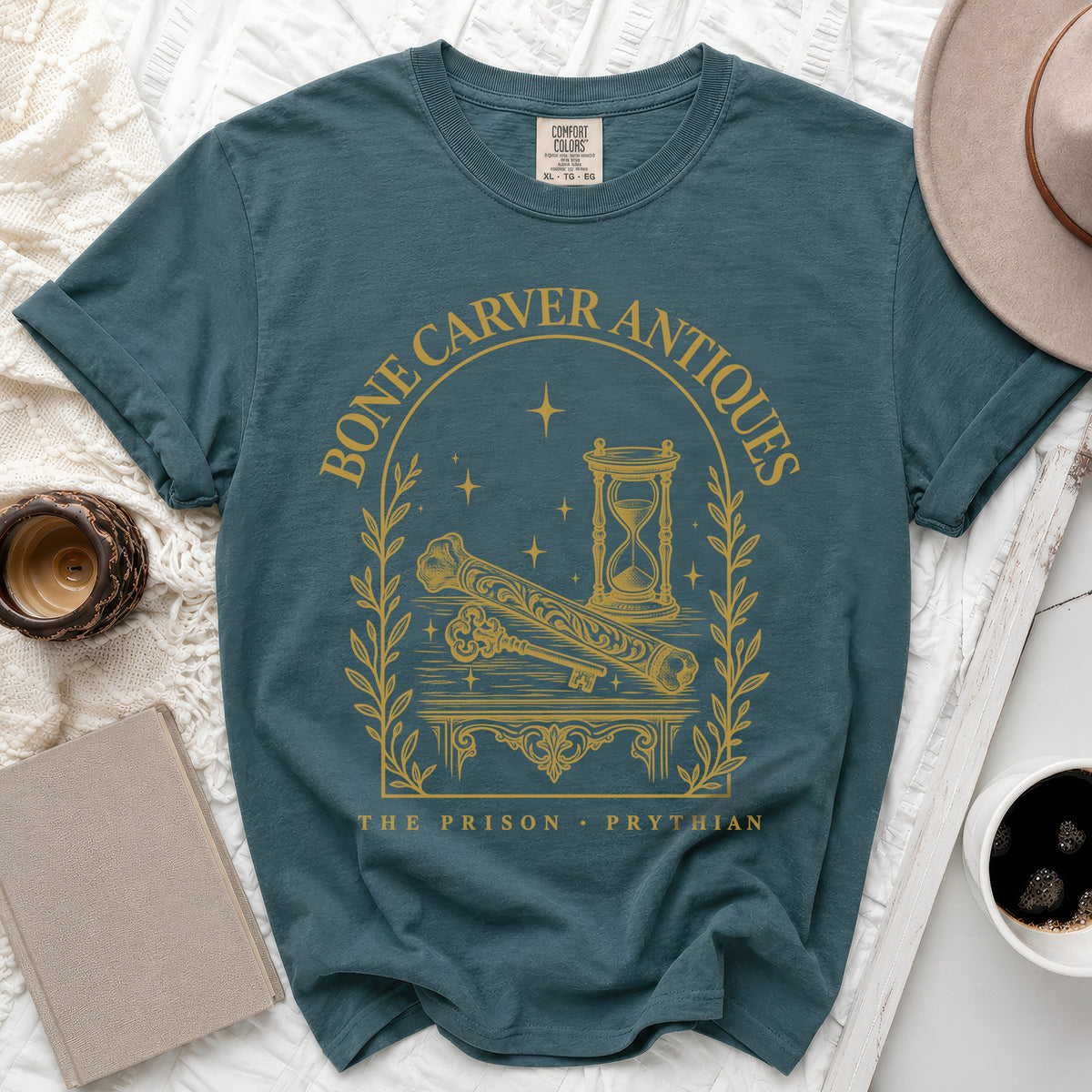Flat lay of a teal t-shirt featuring a golden arched design with bones, hourglass, and text reading Bone Carver Antiques.