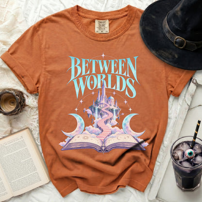 Orange t-shirt with "BETWEEN WORLDS" text, fantasy castle, and open book graphic.