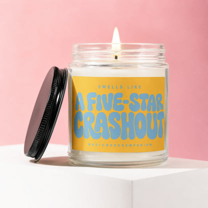 Glass jar candle with lit wick, yellow label reads "SMELLS LIKE A FIVE-STAR CRASHOUT"