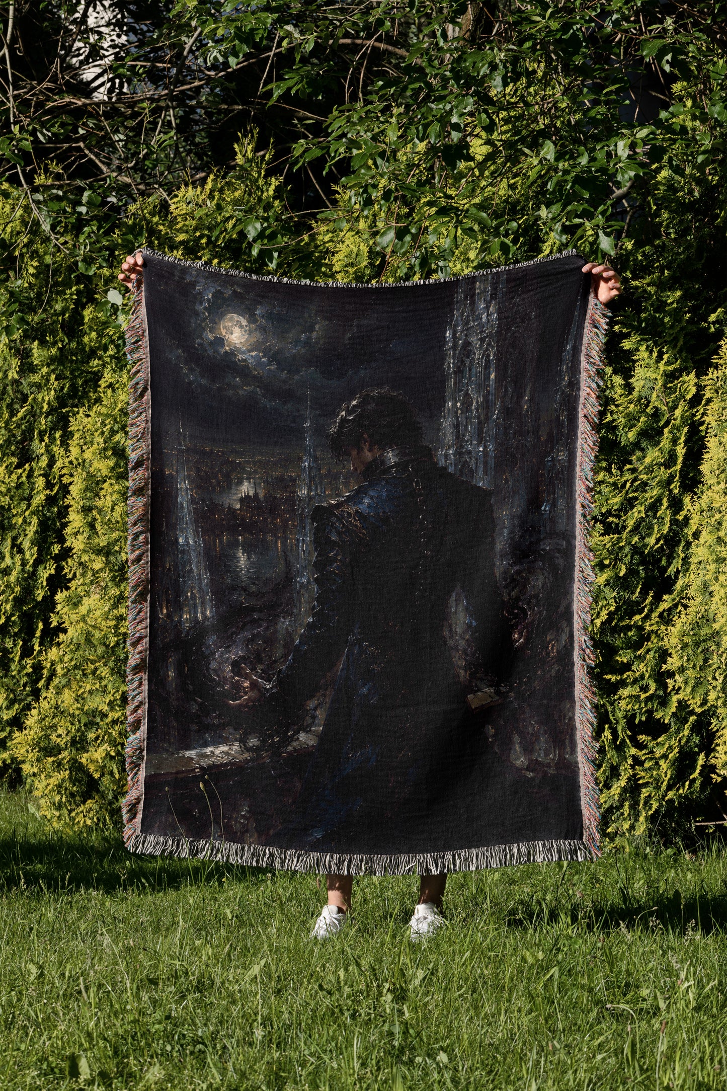 A person is standing in a grassy area, holding up a large black blanket with a painting of a person on it.