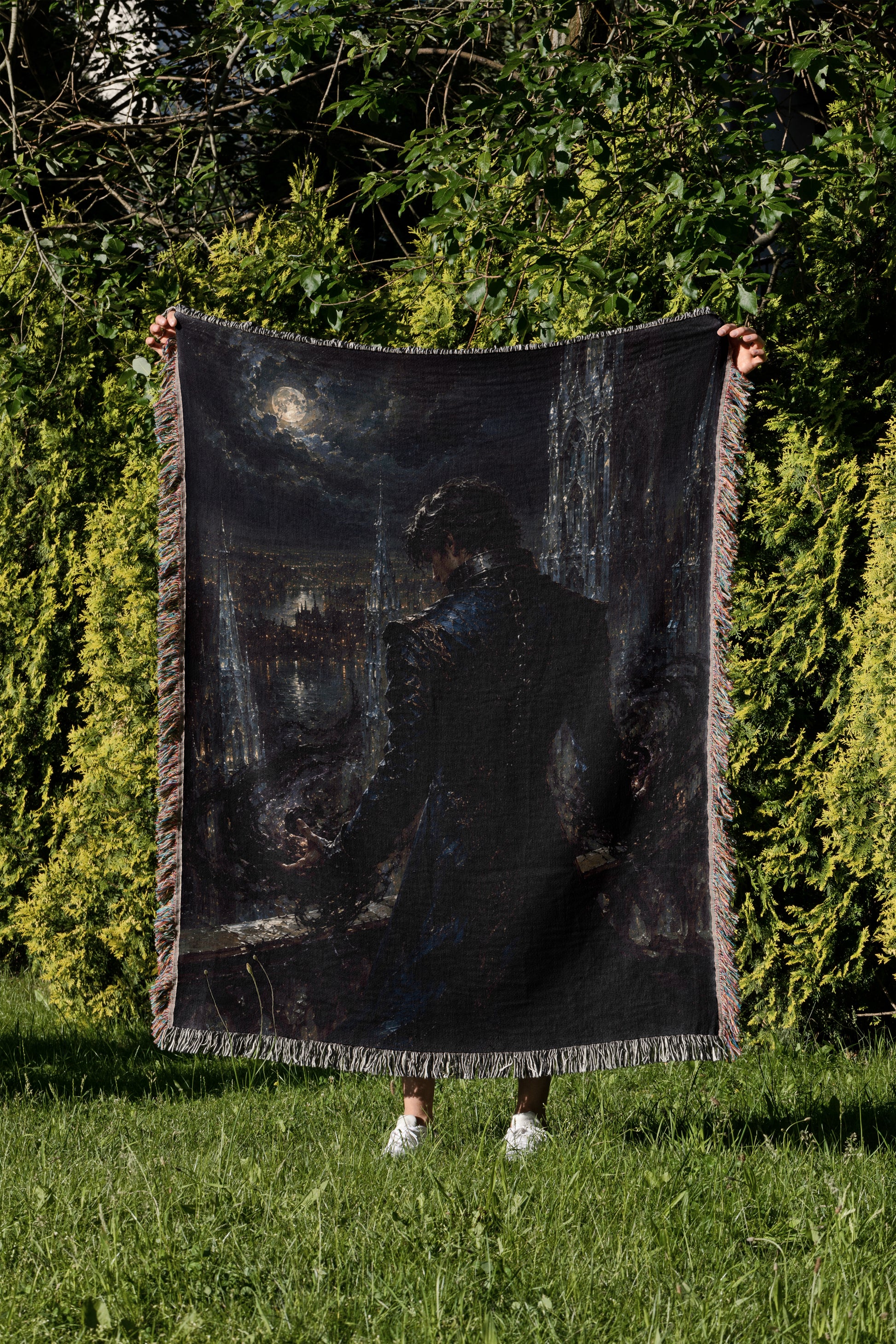 A person is standing in a grassy area, holding up a large black blanket with a painting of a person on it.