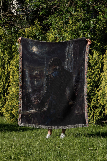 A person is standing in a grassy area, holding up a large black blanket with a painting of a person on it.