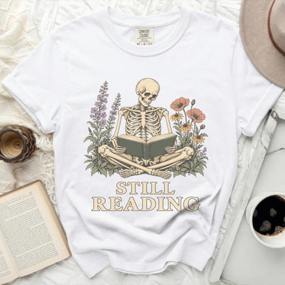 Skeleton reading a book surrounded by flowers with text "STILL READING".