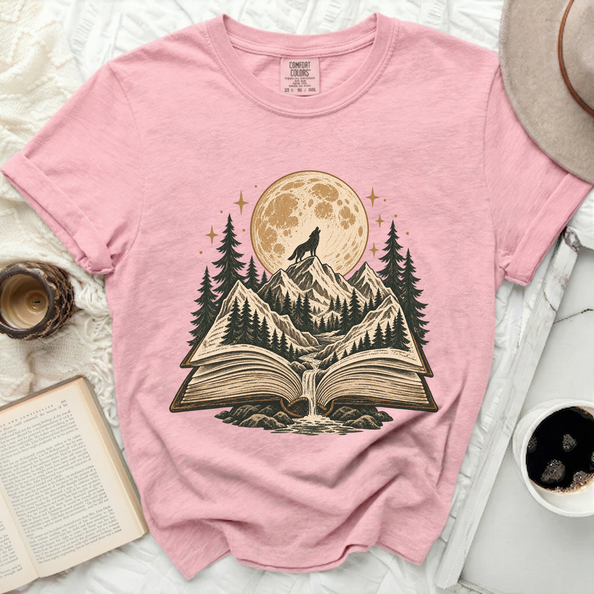 Pink t-shirt with open book, wolf howling at moon, and forest scene design.