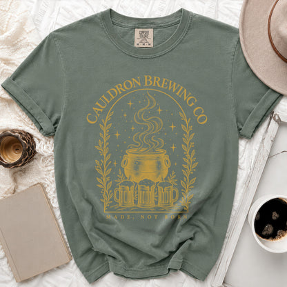 A moss green t-shirt features a yellow graphic of a bubbling cauldron, beer mugs, and stars.