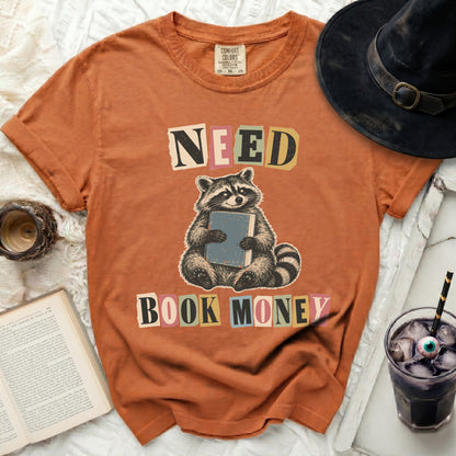 Orange t-shirt with graphic of raccoon holding book, text reads "NEED BOOK MONEY."