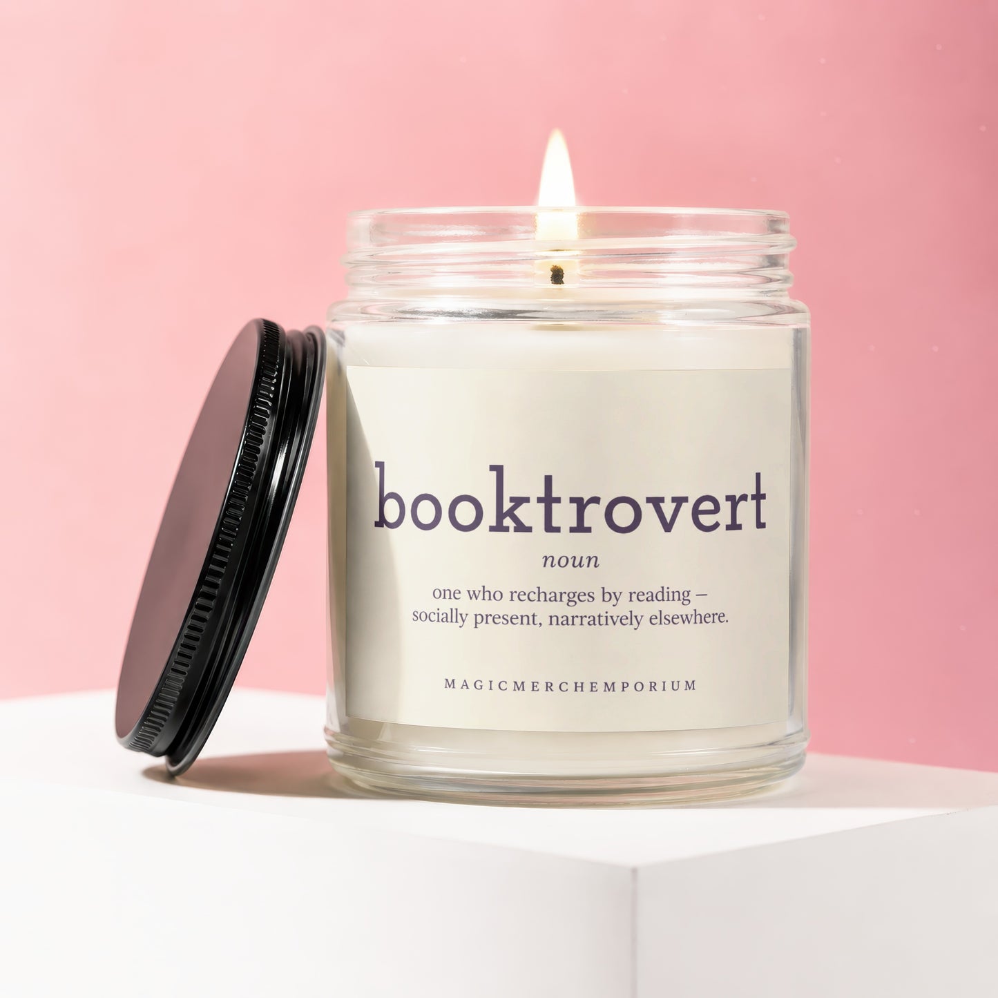 Glass candle with lit wick, black lid, and "booktrovert" label in a pink studio.